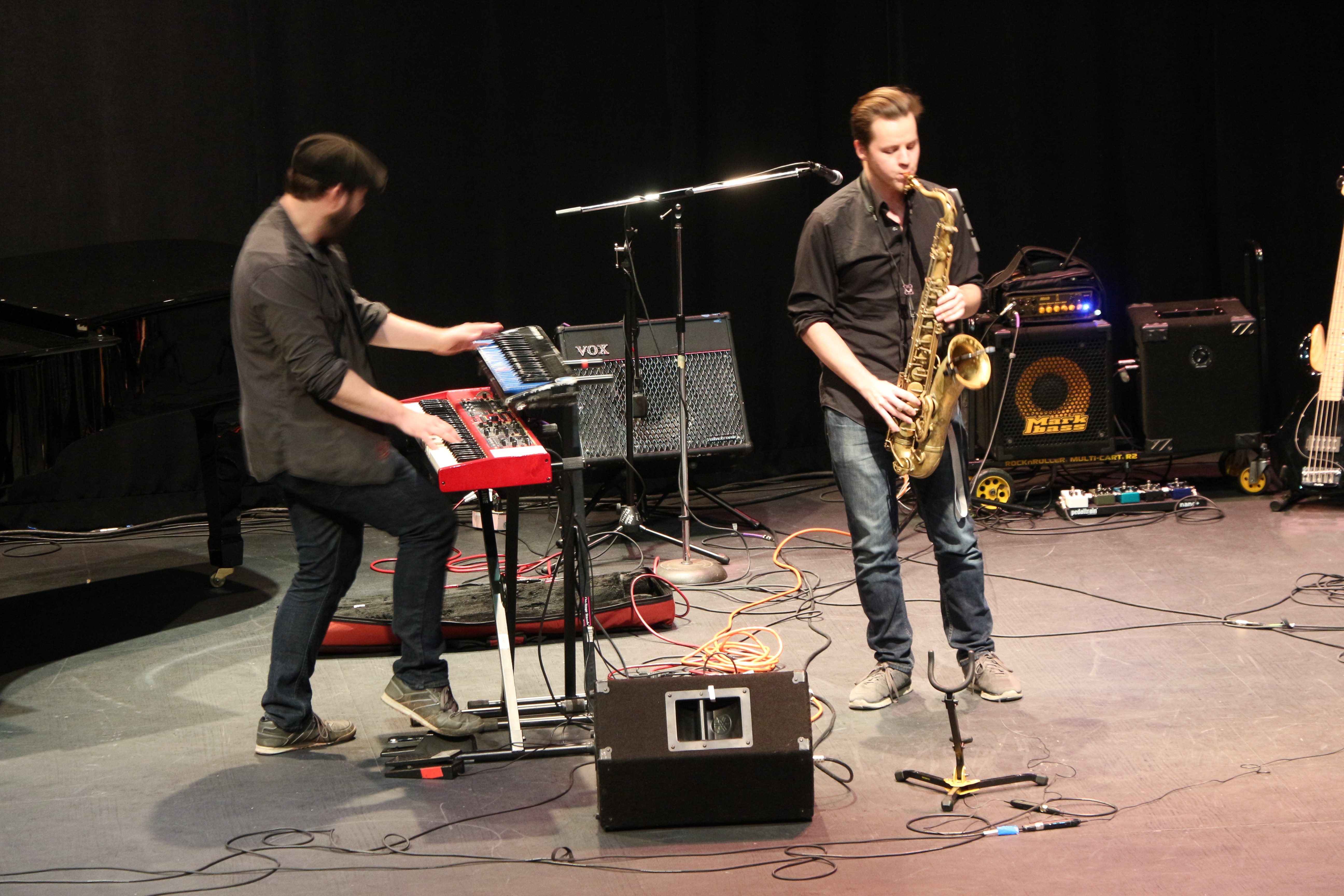 Man wearing black, playing the keyboard on stage with saxophonist in black shirt and blue jeans. 